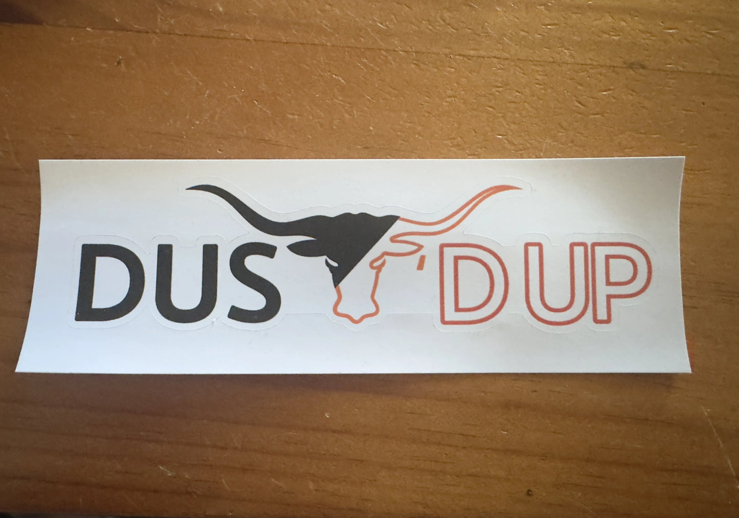 Sticker with DUSt'd up' text on a white background, placed on a wooden surface.